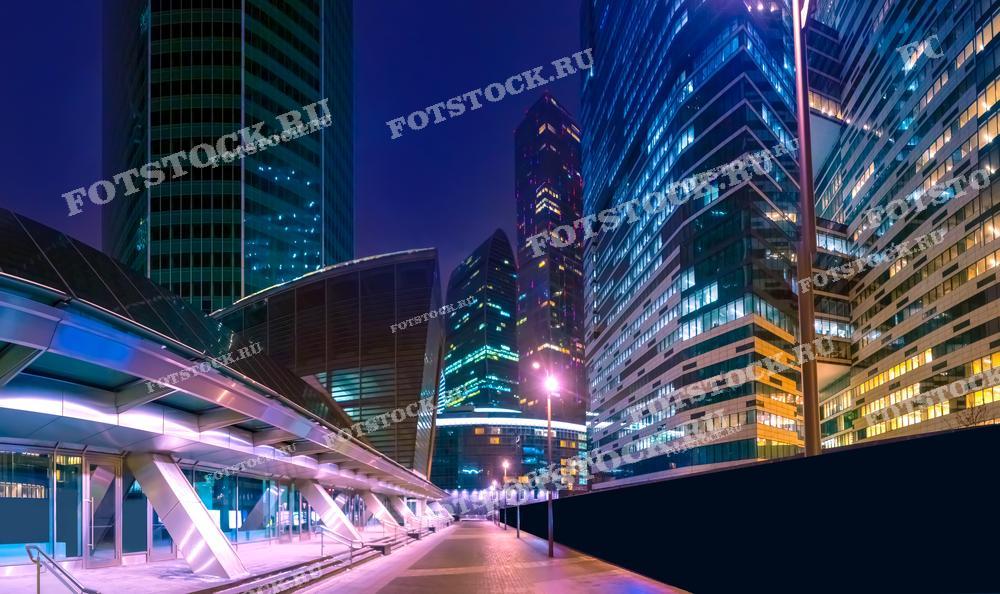 На фотографии изображены небоскребы делового центра Москва-Сити, одного из главных финансовых районов столицы. Высотные здания ярко освещены в ночное время, отражая современные технологии и архитектурные решения. Свет фонарей и иллюминация офисов создают атмосферу урбанистического ландшафта. Пустая улица между небоскребами подчеркивает величие и масштабность этих архитектурных сооружений. Москва-Сити — символ современных достижений столицы в сфере бизнеса и технологий.